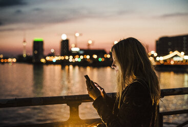 Young woman by waterfront, at dusk, using smartphone - CUF21614