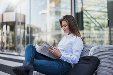 Portrait of young businesswoman sitting on bench reading newspaper - DIGF04565