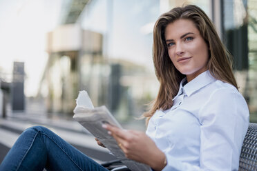 Portrait of smiling young businesswoman sitting on bench with newspaper - DIGF04568