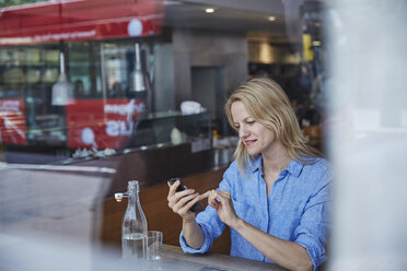 Ältere Frau sitzt in einem Café und benutzt ihr Smartphone, der Bus spiegelt sich im Fenster - CUF21667