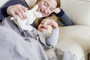 Baby girl and mother playing with toy rabbit on sofa - CUF21711