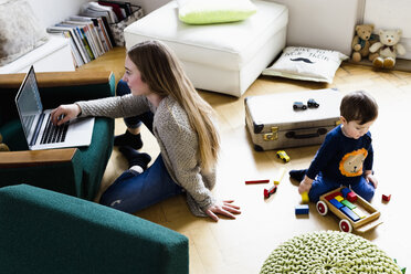 Mittlere erwachsene Frau tippt auf einem Laptop, während ihr kleiner Sohn auf dem Boden spielt - CUF21734