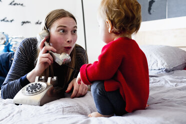 Kleines Mädchen und Mutter auf dem Bett, die am Festnetztelefon ihr Gesicht verziehen - CUF21757