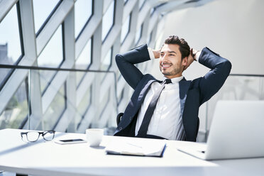 Smiling businessman leaning back at desk in modern office - BSZF00482