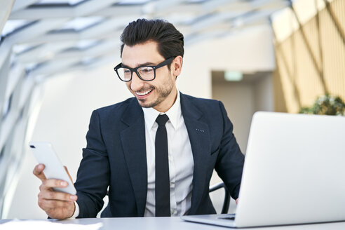 Lächelnder Geschäftsmann, der auf sein Handy am Schreibtisch in einem modernen Büro schaut - BSZF00485