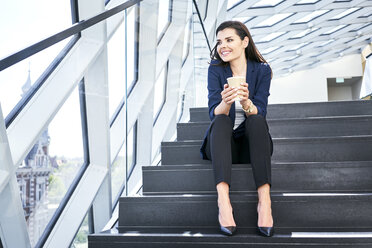 Lächelnde Geschäftsfrau sitzt auf einer Treppe und macht eine Kaffeepause in einem modernen Büro - BSZF00548