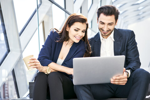 Lächelnde Geschäftsfrau und Geschäftsmann teilen sich einen Laptop auf einer Treppe in einem modernen Büro, lizenzfreies Stockfoto