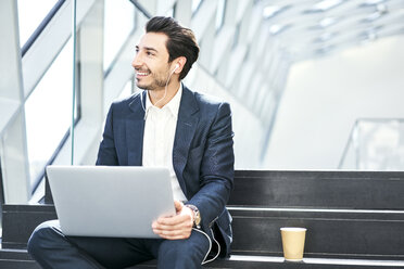 Smiling businessman sitting on stairs wearing earphones and using laptop - BSZF00554