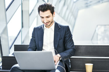 Lächelnder Geschäftsmann, der auf einer Treppe sitzt, Kopfhörer trägt und einen Laptop benutzt - BSZF00555