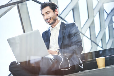 Smiling businessman sitting on stairs wearing earphones and using laptop - BSZF00556