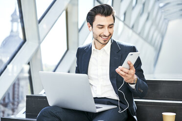 Lächelnder Geschäftsmann, der auf einer Treppe sitzt, Kopfhörer trägt und ein Handy und einen Laptop benutzt - BSZF00558