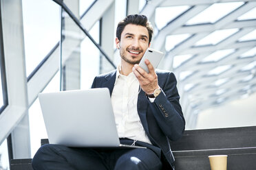 Smiling businessman sitting on stairs wearing earphones using cell phone and laptop - BSZF00559