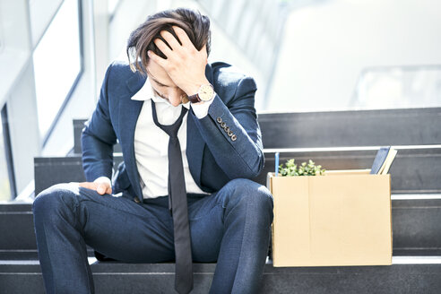 Depressed businessman sitting on stairs with belongings in cardboard box beside him - BSZF00560