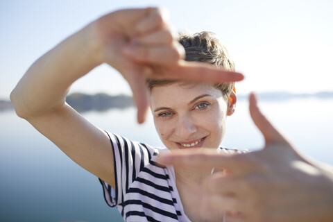 Porträt einer Frau vor einem See, die mit ihren Fingern einen Rahmen formt, lizenzfreies Stockfoto