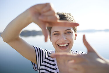 Portrait of laughing woman in front of lake shaping frame with her fingers - PNEF00652