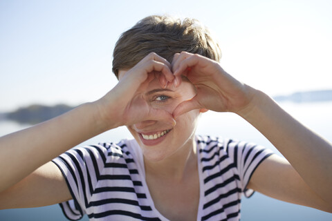 Porträt einer lächelnden Frau vor einem See, die mit ihren Fingern ein Herz formt, lizenzfreies Stockfoto