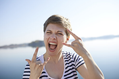 Porträt einer schreienden Frau vor einem See mit Rock-and-Roll-Zeichen - PNEF00654