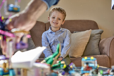 Lächelnder Junge sitzt auf der Couch und betrachtet Spielzeug auf dem Tisch - BEF00139