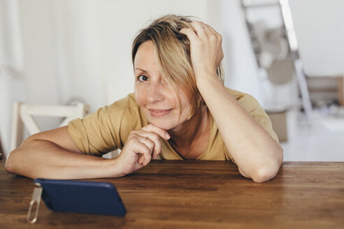 Porträt einer lächelnden Frau, die mit einem Smartphone am Tisch sitzt, in einer neuen Wohnung - KMKF00256