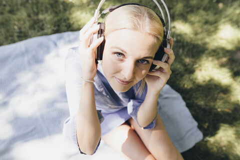 Porträt einer lächelnden jungen Frau, die auf einer Decke sitzt und Kopfhörer trägt, lizenzfreies Stockfoto