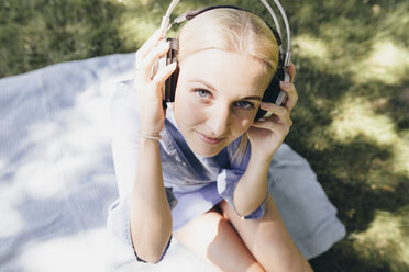 Porträt einer lächelnden jungen Frau, die auf einer Decke sitzt und Kopfhörer trägt - KMKF00277