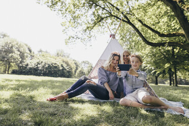 Two young women and a boy taking a selfie next to teepee in a park - KMKF00280