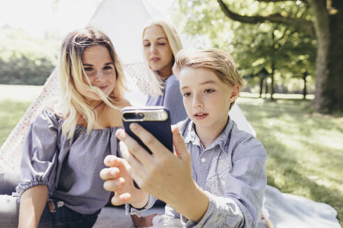 Zwei junge Frauen und ein Junge machen ein Selfie neben einem Tipi in einem Park - KMKF00281