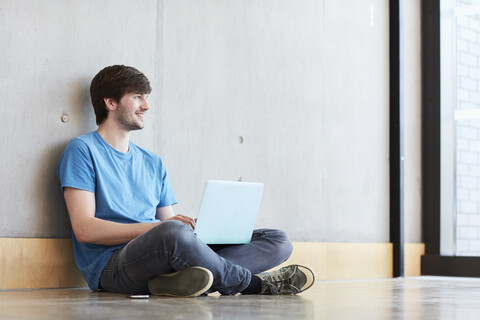 Junger männlicher Student mit Laptop auf dem Boden sitzend in einer Hochschuleinrichtung, lizenzfreies Stockfoto