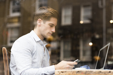 Blick aus dem Fenster auf einen jungen Geschäftsmann, der in einem Café Texte auf seinem Smartphone liest - CUF22261