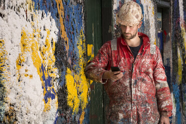 Männlicher Schiffsmaler liest Smartphone-Text und lehnt sich an eine mit Farbe bespritzte Wand - CUF22397