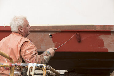 Älterer männlicher Schiffsmaler, der den Schiffsrumpf rot anstreicht, in einer Werft für Schiffsmaler - CUF22415