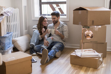 Moving house: Young couple sitting in room full of boxes, drinking hot drink - ISF07904