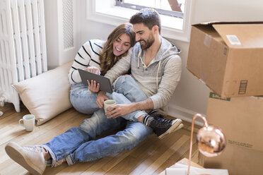 Moving house: Young couple sitting in room full of boxes, looking at digital tablet - ISF07905