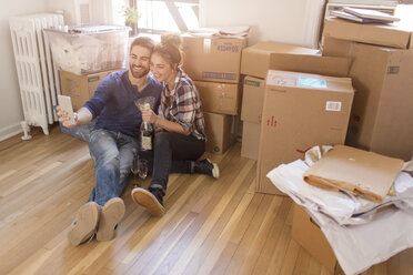 Moving house: Young couple sitting in room full of boxes, holding champagne bottle, taking self portrait with smartphone - ISF07912