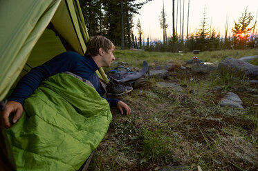 Männlicher Camper im Schlafsack beobachtet den Sonnenuntergang auf dem Midnight Ridge, Colville National Forest, Washington State, USA - ISF08009