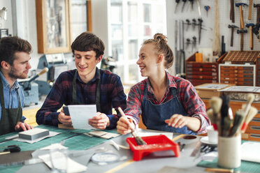 Junge Handwerkerin hält Pinsel und lacht und lächelt mit zwei jungen Handwerkern in einem kreativen Druckstudio - CUF22543