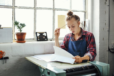 Junge Handwerkerin am Fenster, die sich an eine Buchdruckmaschine lehnt und Papierkram im Atelier für Buchkunst betrachtet - CUF22548