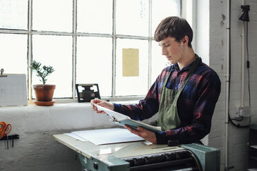 Junger Handwerker am Fenster, der sich an eine Buchdruckmaschine lehnt und auf ein Klemmbrett im Atelier für Buchkunst schaut - CUF22549