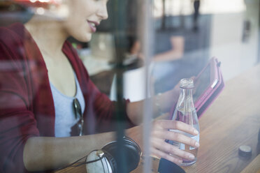 Junge Frau sitzt in einem Café, hält eine Flasche Wasser in der Hand und benutzt ein digitales Tablet - CUF22612