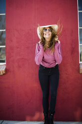 Woman in front of red wall wearing sunhat and sunglasses smiling - ISF08242