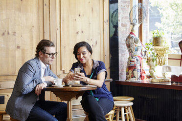 Mann und Frau sitzen in einem Café, trinken Kaffee und schauen auf ihr Smartphone - ISF08384