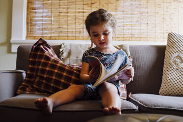 Weibliches Kleinkind sitzt auf dem Sofa und liest ein Bilderbuch - ISF08724