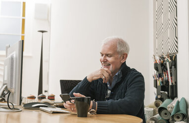 Senior man smiling, using mobile phone at work desk - CUF22806