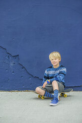 Porträt eines Jungen, der auf einem Skateboard vor einer blauen Wand sitzt - ISF08746