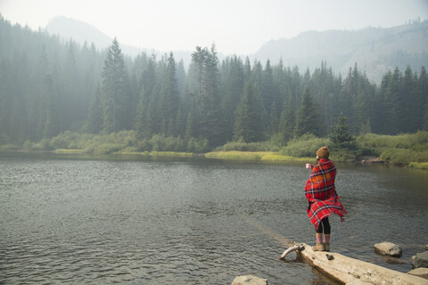 Frau in Schottenkaro-Decke eingewickelt, Kaffee trinkend am nebligen See, Mount Hood National Forest, Oregon, USA, lizenzfreies Stockfoto