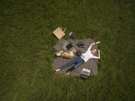 Bird's eye view of woman lying on blanket on meadow with dog and utensils - ONF01124