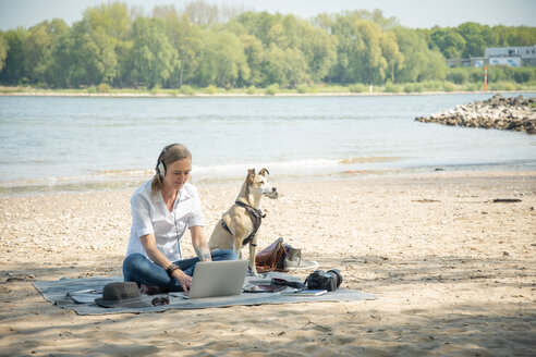 Frau sitzt auf einer Decke an einem Fluss mit Hund, trägt Kopfhörer und benutzt einen Laptop - ONF01126