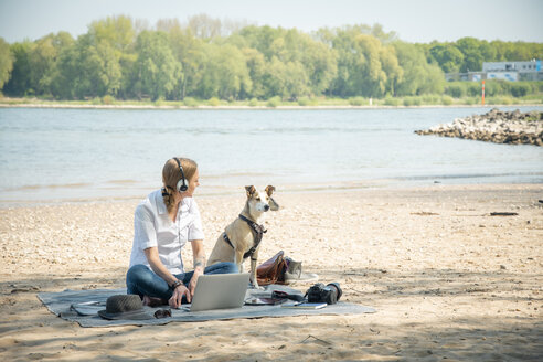 Woman sitting on blanket at a river with dog wearing headphones and using laptop - ONF01127