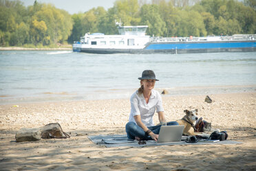 Smiling woman sitting on blanket at a river with dog using laptop - ONF01128