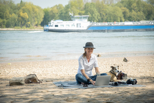 Lächelnde Frau sitzt auf einer Decke an einem Fluss mit Hund und benutzt einen Laptop - ONF01128